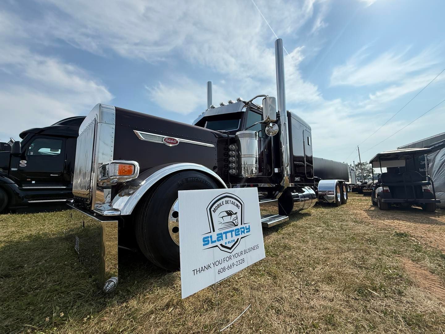A custom, dark-painted semi-truck sits on grass under a blue sky, partially obscured by a Slattery advertising sign.