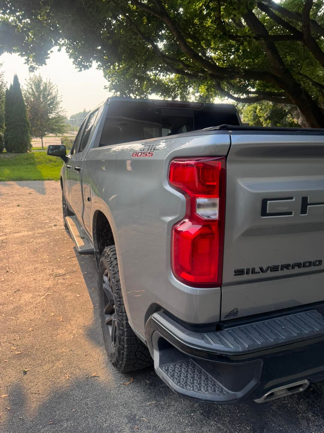 A silver Chevrolet Silverado pickup truck parked on a paved driveway near a large tree at sunset.