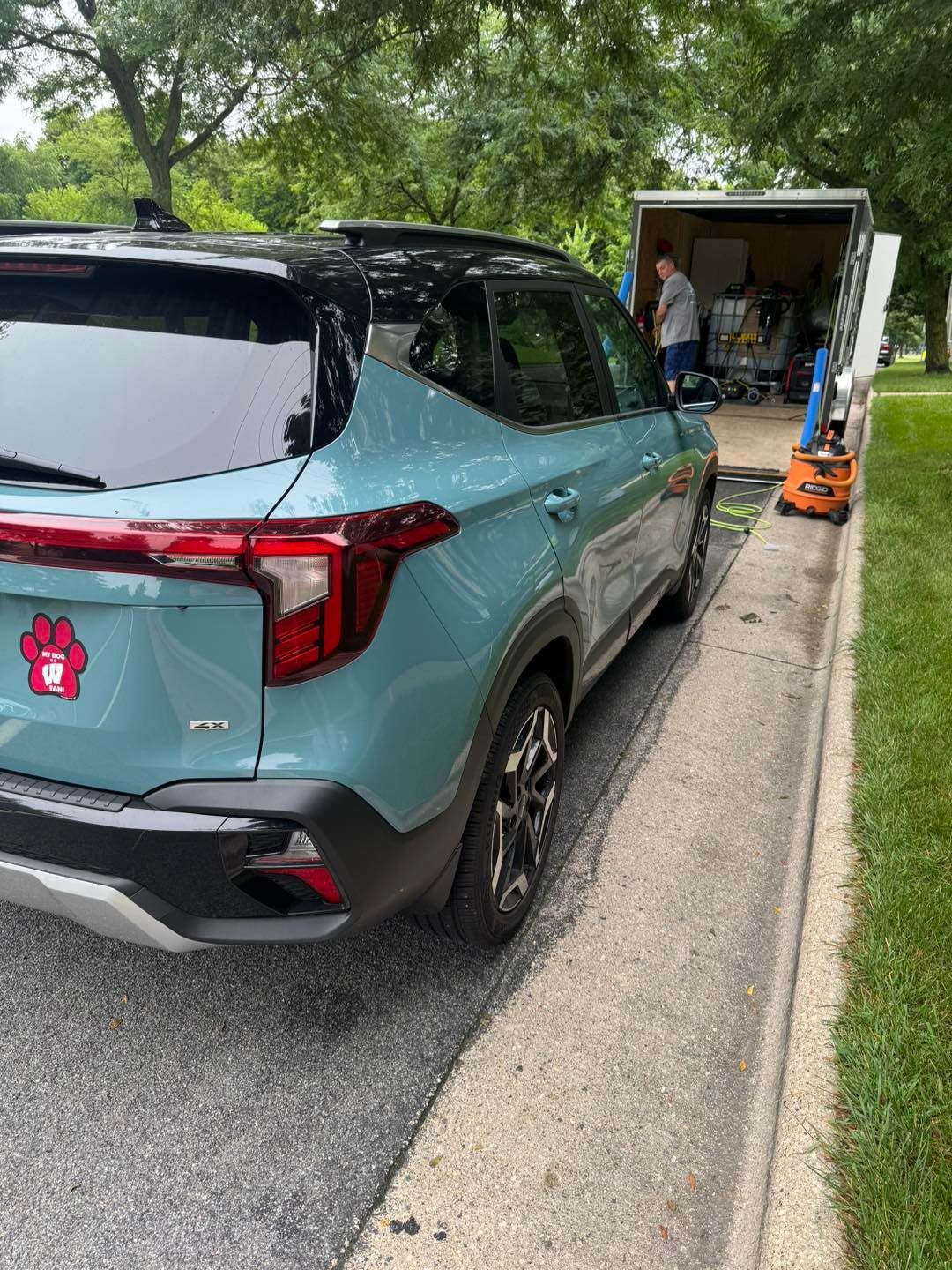 Teal SUV parked on a street near a trailer being loaded with equipment by a person.