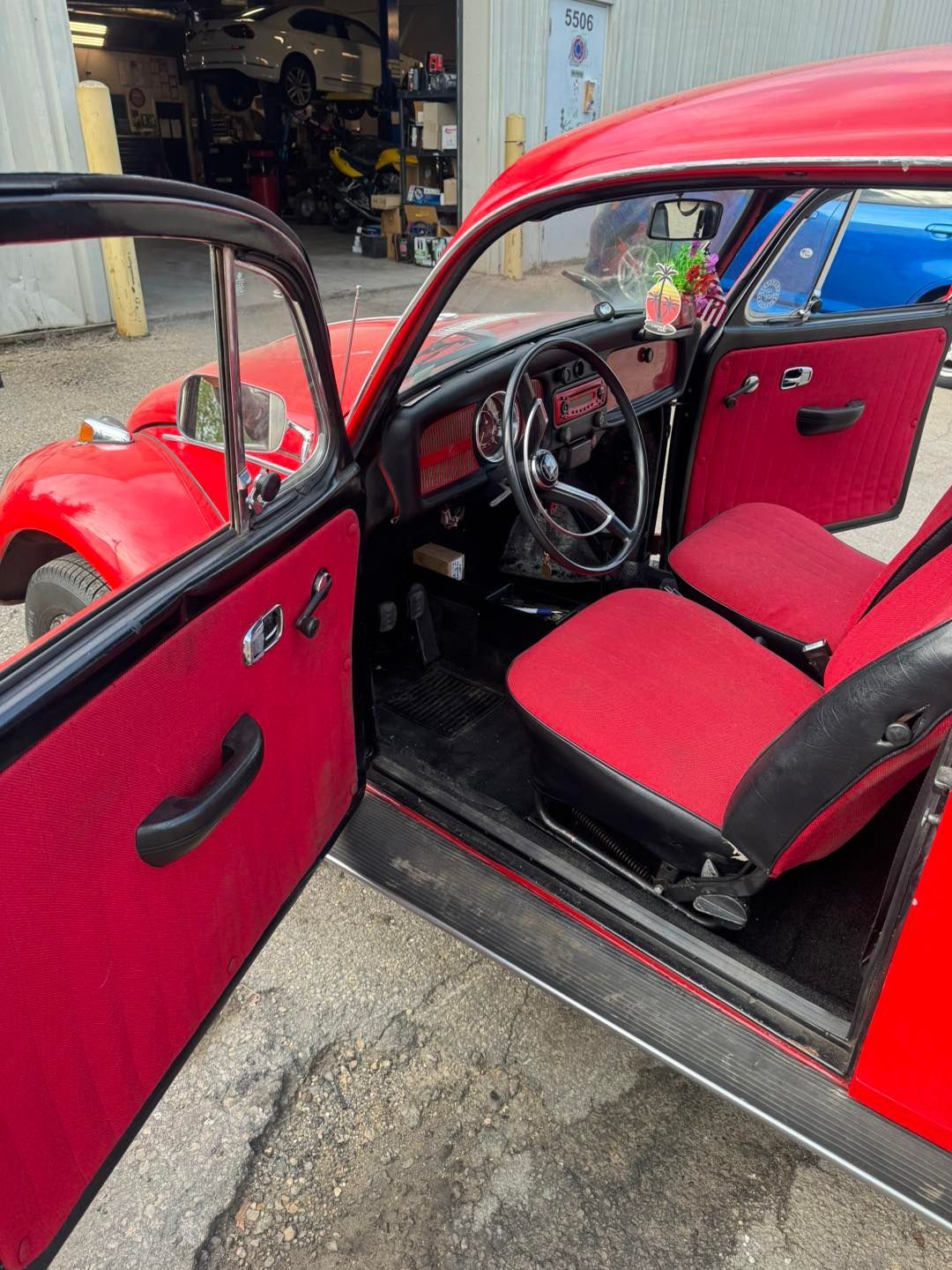 The interior of a red vintage Volkswagen Beetle with matching red upholstery, seen through the open driver-side door.