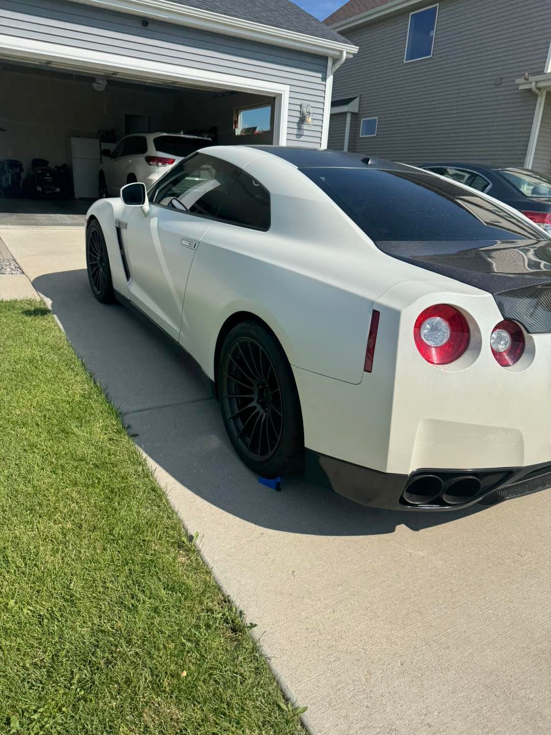 A white Nissan GT-R sports car with dark wheels and a carbon fiber trunk parked on a concrete driveway near a garage.