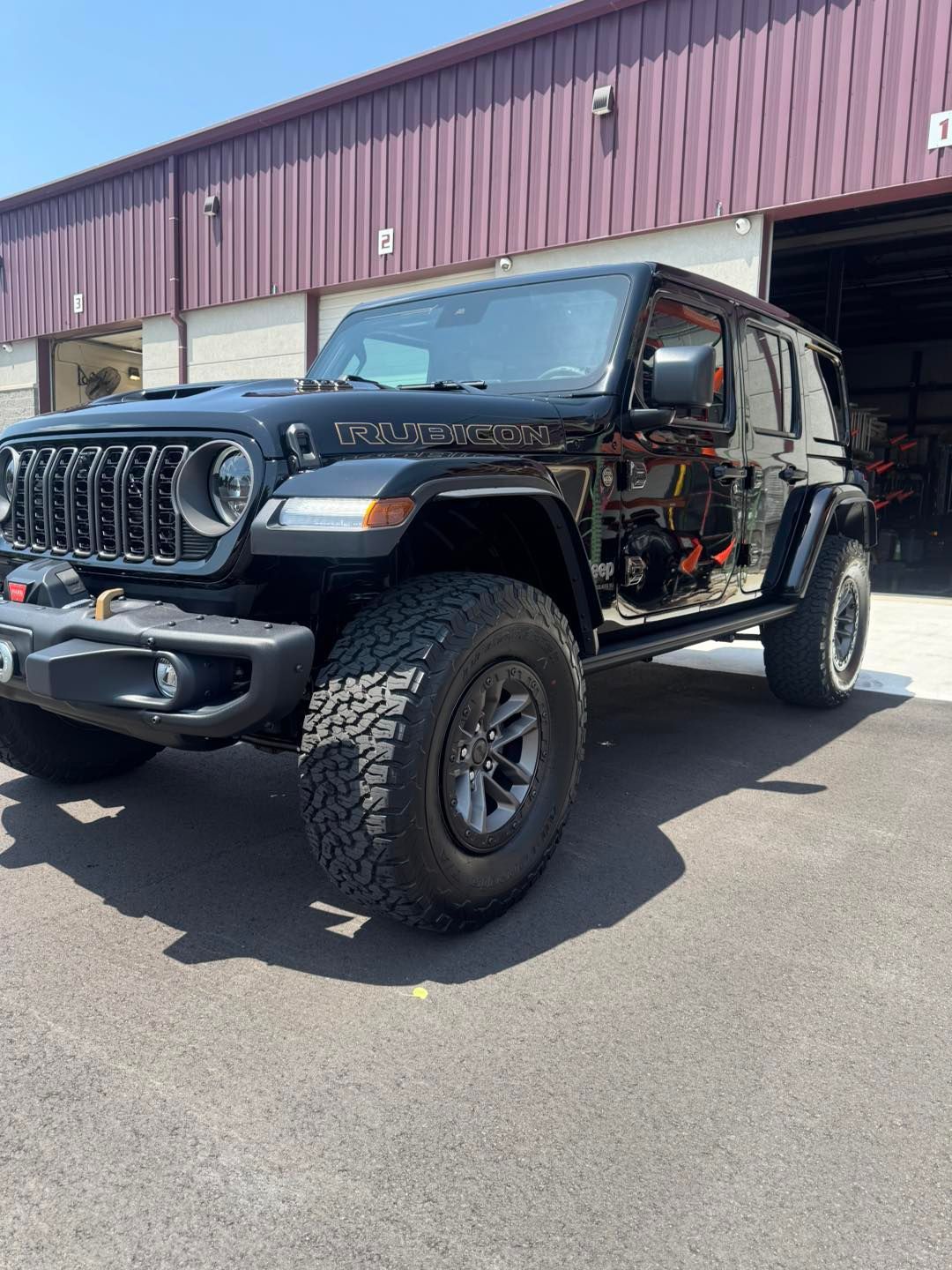 A black Jeep Rubicon parked on an asphalt lot in front of a building with dark red corrugated metal siding.