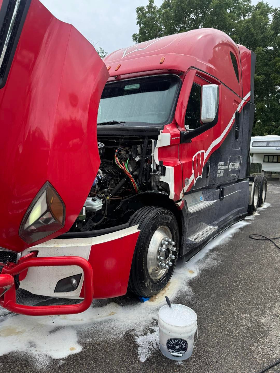A red and black semi-truck with its hood open, being washed with soap on an asphalt surface.