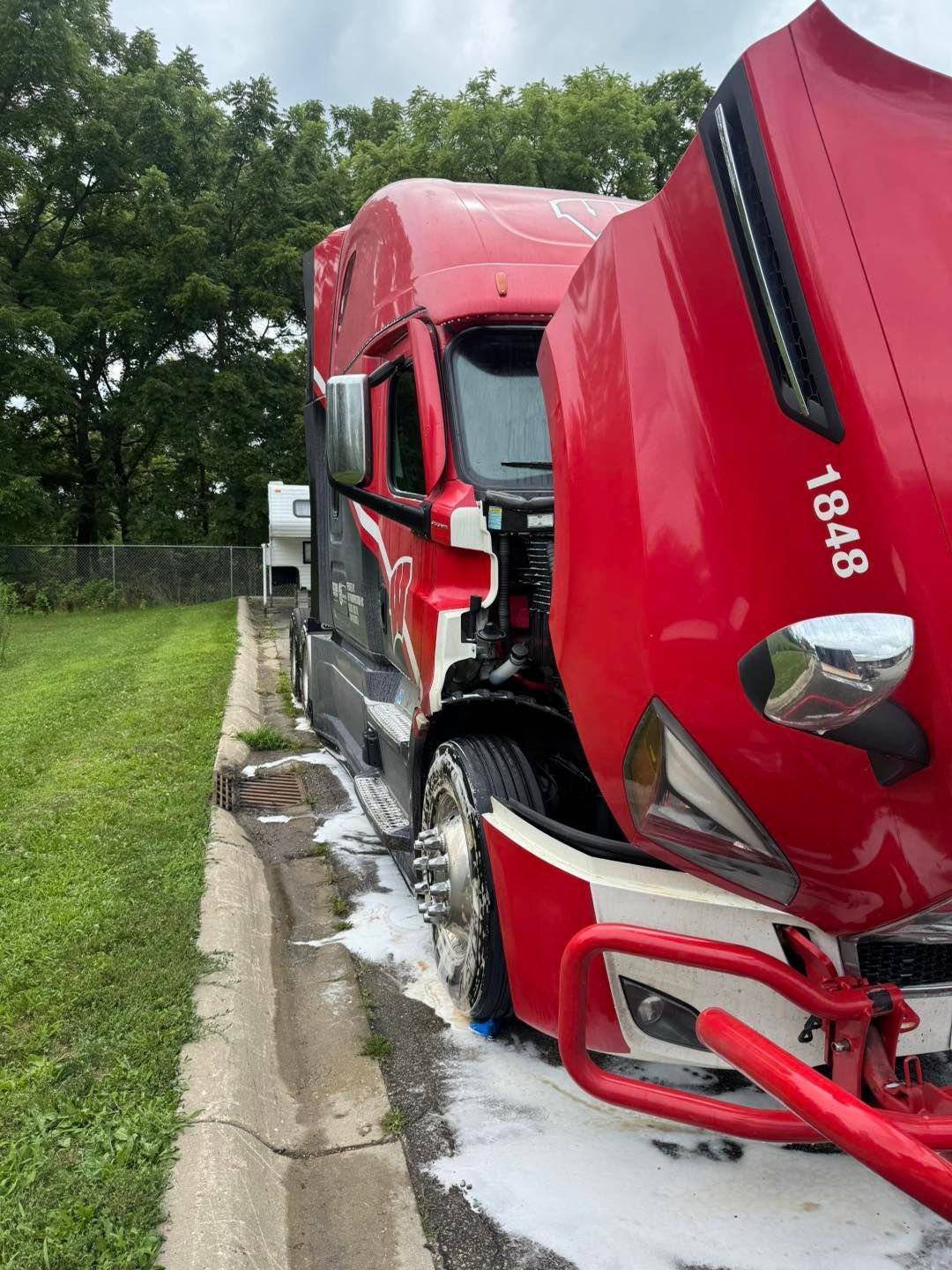 A red semi-truck with its hood open and white fluid leaking onto the pavement beside a grassy area.
