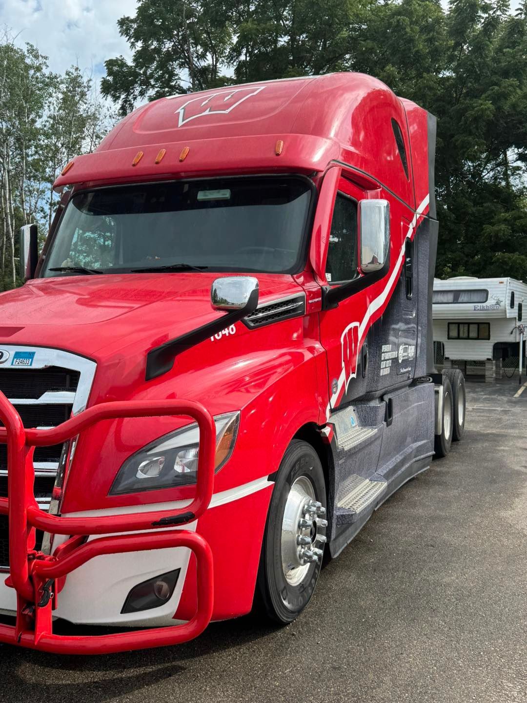 A red and gray semi-truck with a front-mounted brush guard parked in a lot next to a trailer.