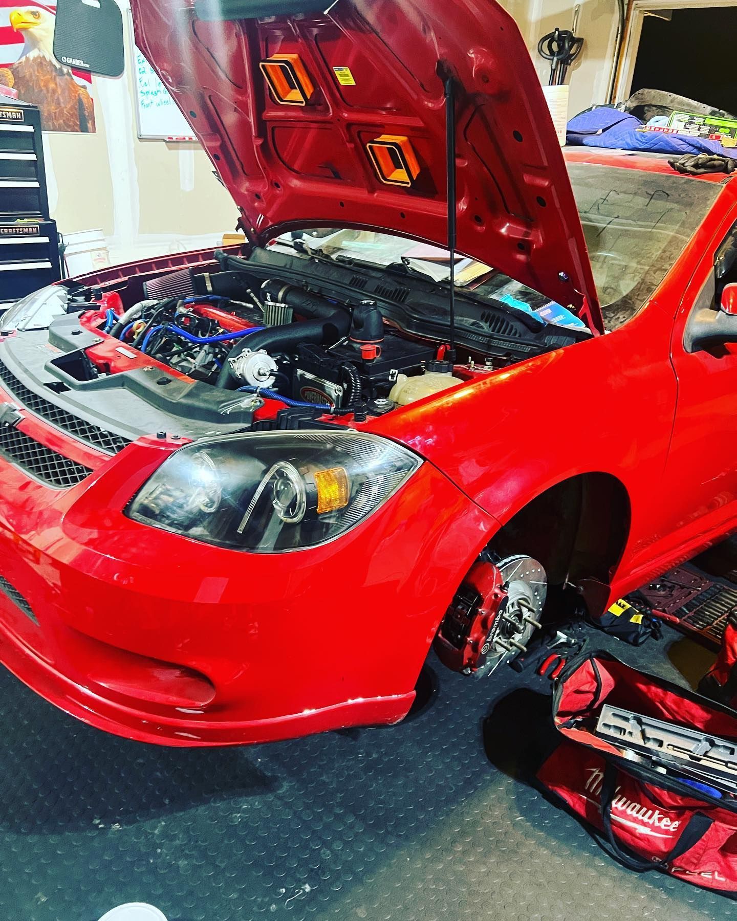 A red car in a garage with its hood open, showing the engine bay and a tool bag sitting by the front wheel assembly.