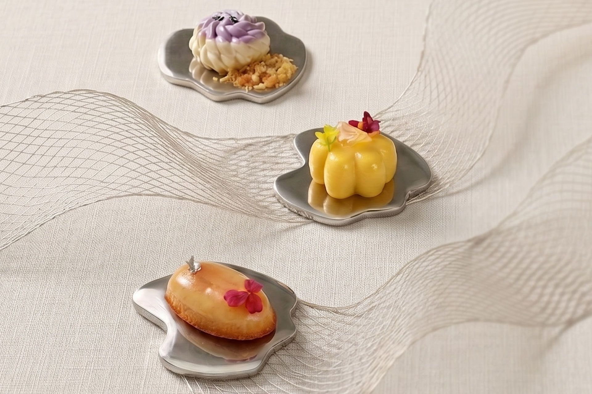 Three small plated pastries on a white textured cloth, each with colorful frosting and garnish