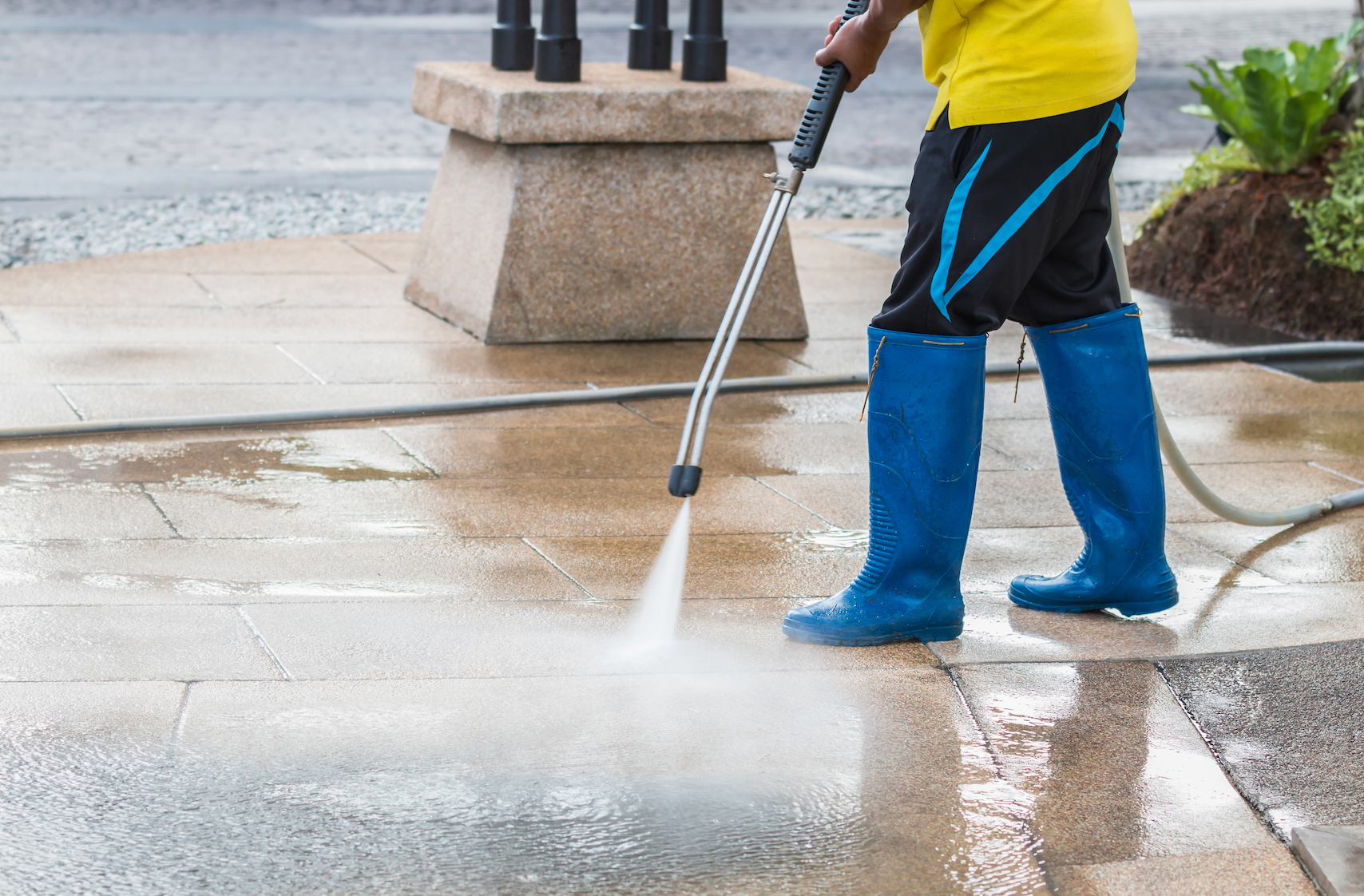A man in blue boots is using a high pressure washer to clean a sidewalk.