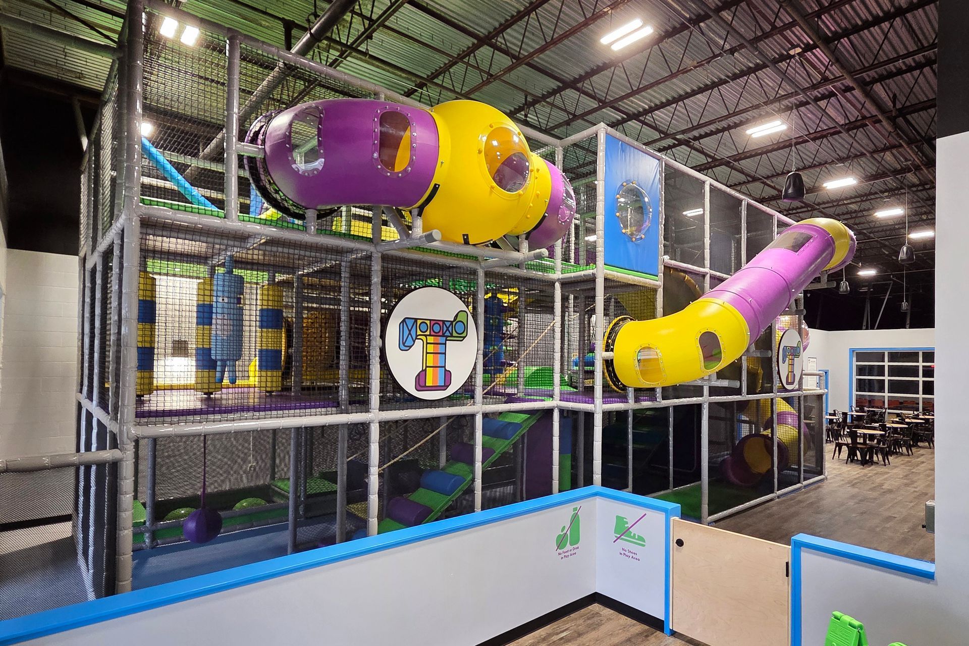 Children in a gymnasium gather around a green mat, with a teacher, interacting and observing.