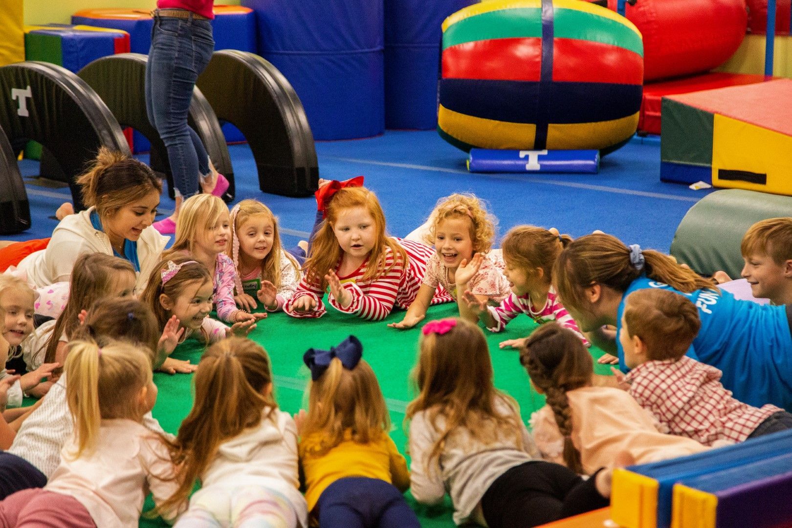 Children in a gymnasium gather around a green mat, with a teacher, interacting and observing.