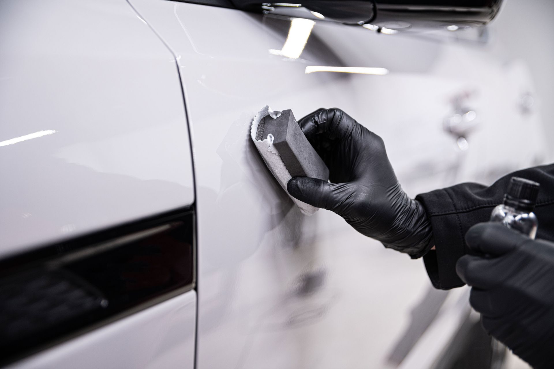Gloved hands applying ceramic coating to a white car panel with an applicator sponge and spray bottle.