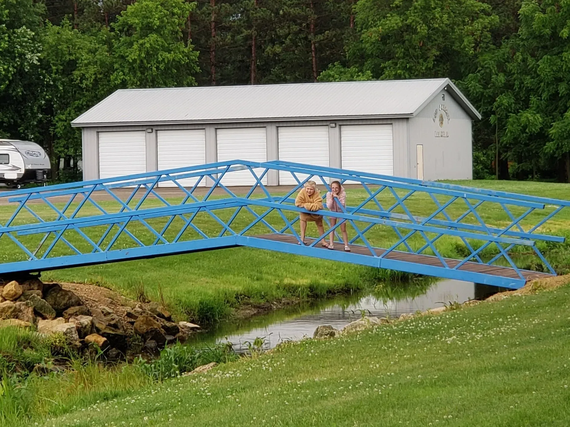 Two people on a blue bridge crossing a creek. A garage is in the background with a camper parked nearby