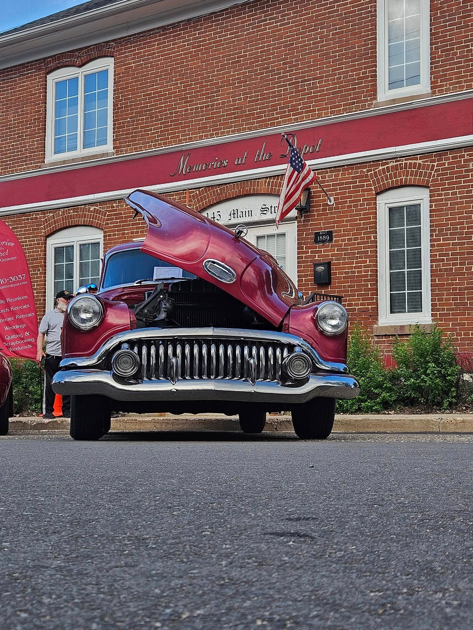 Red classic car with hood up parked on a street in front of a brick building with windows and an American flag.