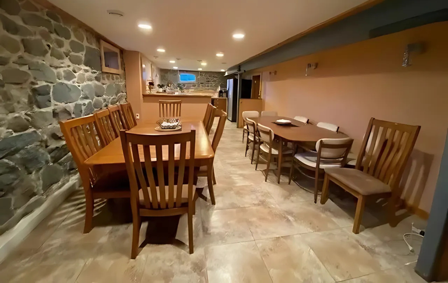 Spacious dining area with two wooden tables and chairs, stone wall, tan flooring, and a small bar in the background.