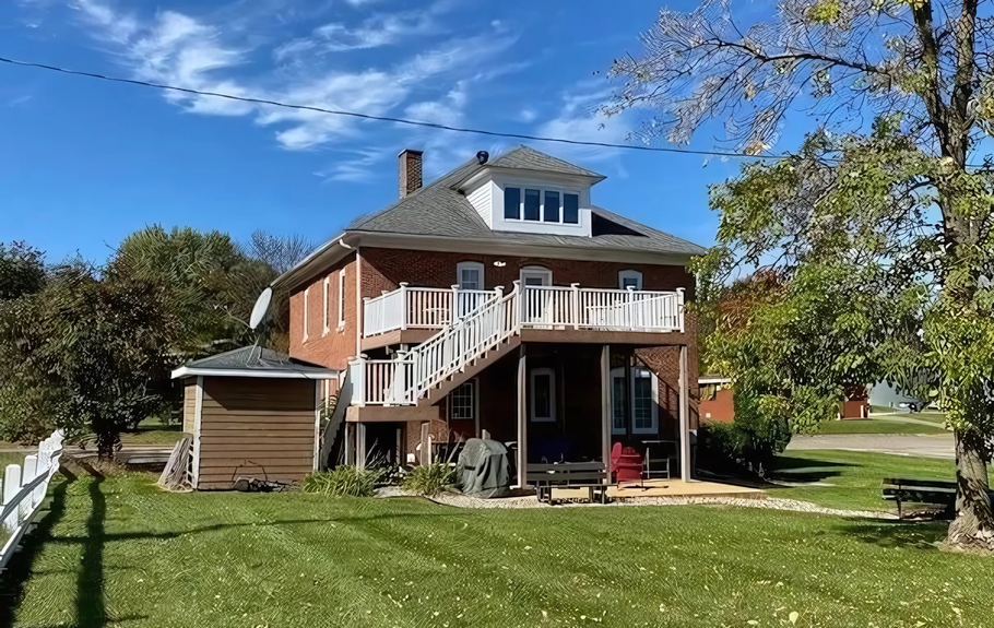 Two-story brick house with a second-story balcony and outdoor staircase, set in a grassy yard on a sunny day.