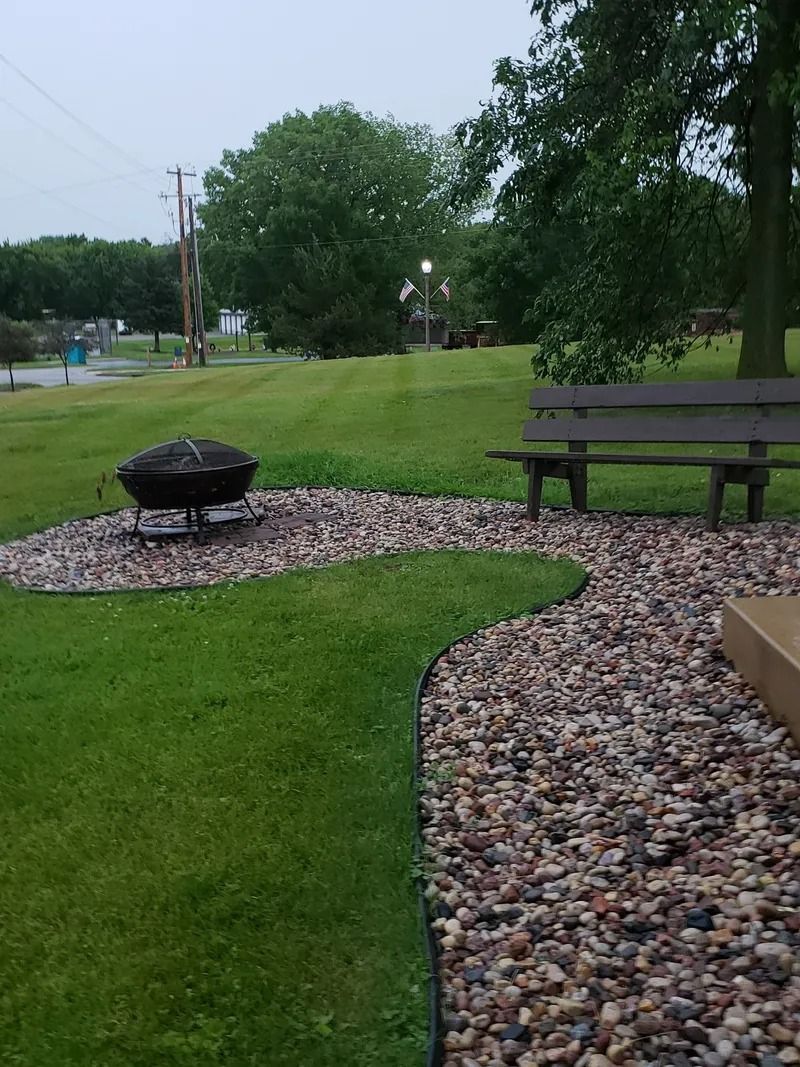 A park scene with a fire pit, bench, and winding pebble path on green grass, under a cloudy sky.