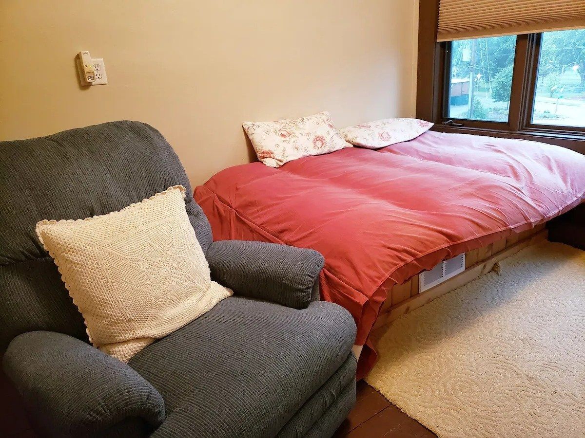 Cozy bedroom with a bed, recliner chair, and a window with a view. Red bedding, a beige chair, and light-colored rug.