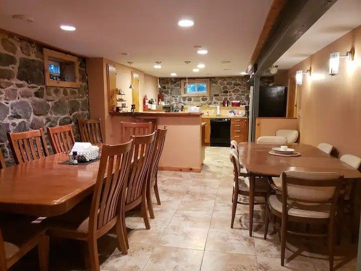 A large dining room with two long wooden tables, stone walls, and a kitchen area in the background.