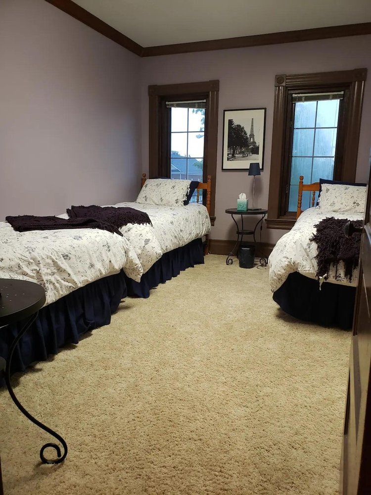 Two twin beds in a room with lavender walls, dark wood trim, and floral bedding. A small table stands between two windows.