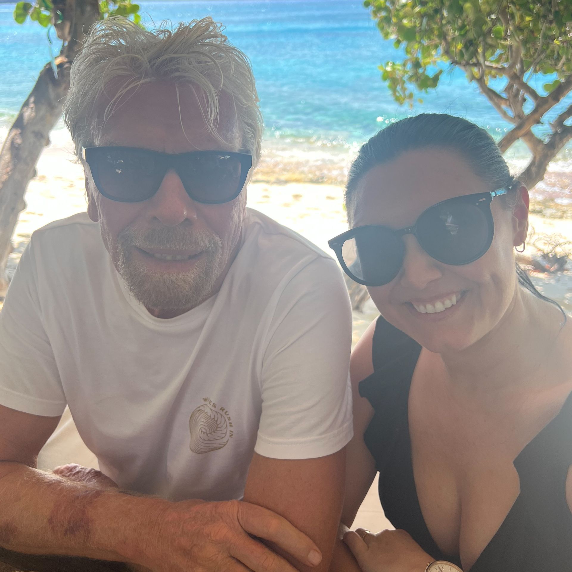 A man and a woman wearing sunglasses are sitting next to each other on a beach.