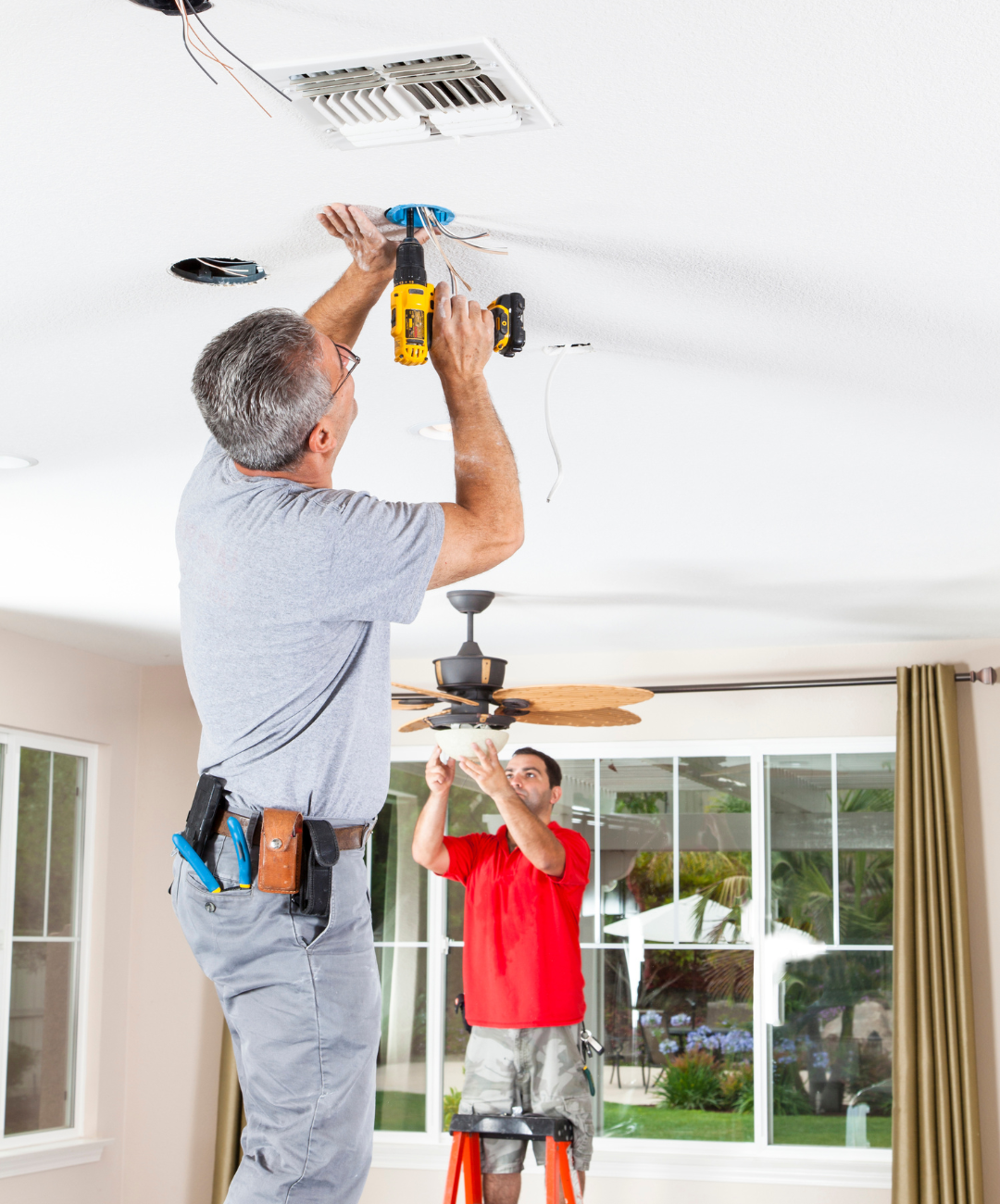 Two men are working on a ceiling fan in a living room.