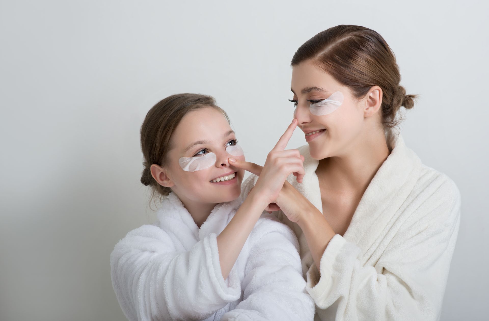 Two people wearing white robes and eye patches playfully touch each other's noses against a plain background.