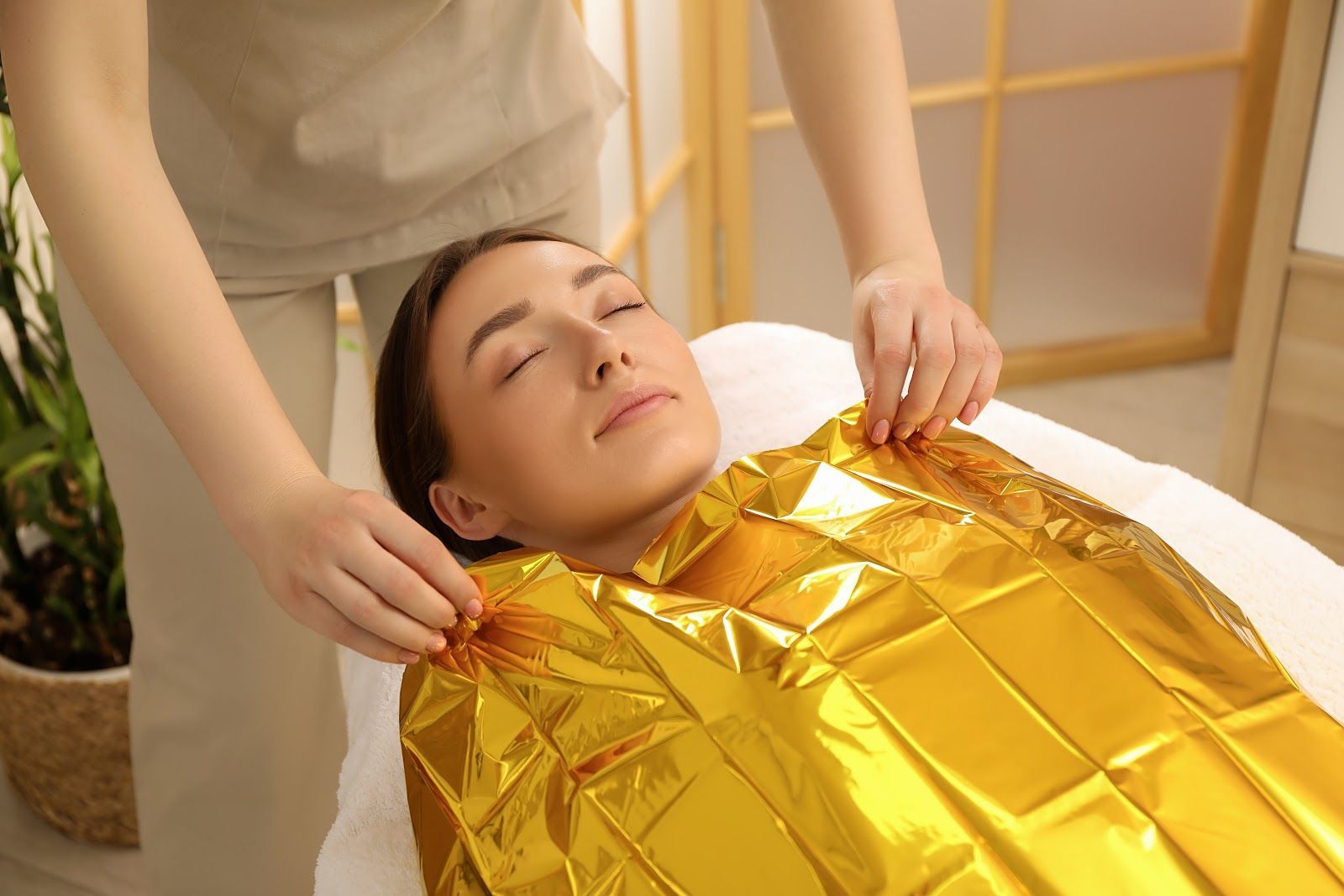 Woman relaxing on a massage table, wrapped in a gold blanket, while someone adjusts it.