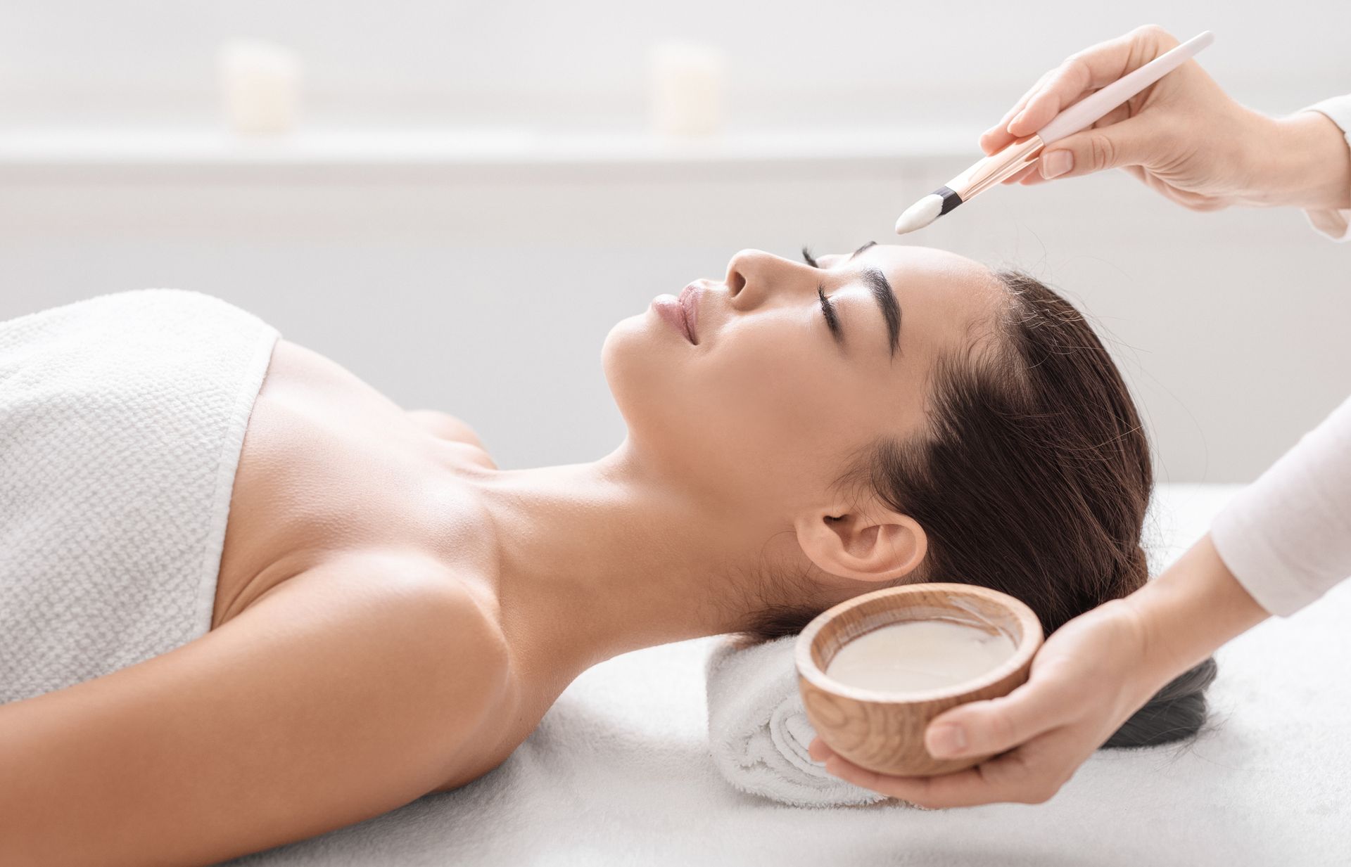A person receives a facial treatment with a brush while lying on a spa bed, with a technician holding a small bowl nearby.