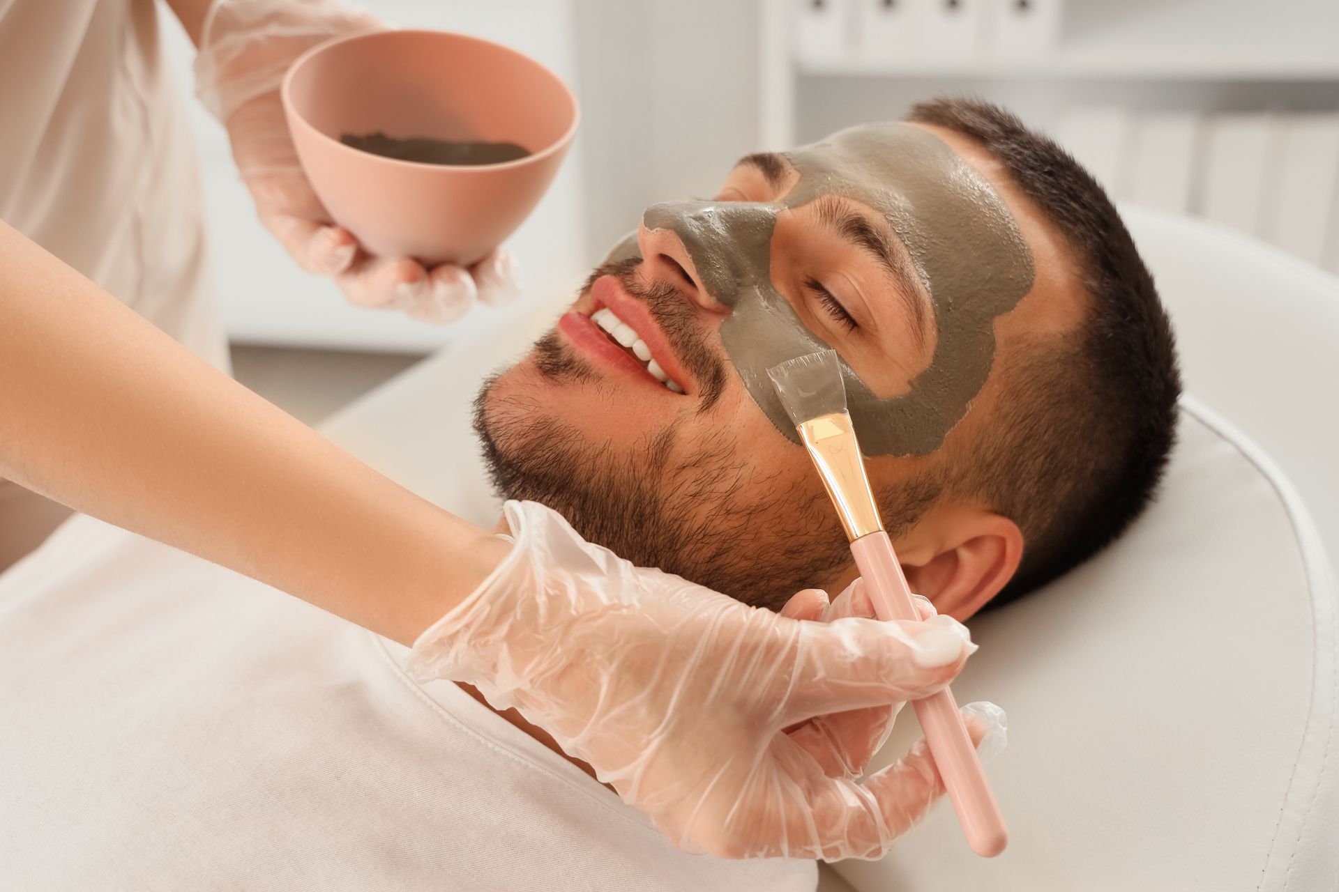 A person receives a facial treatment with a grey clay mask applied by a professional wearing gloves in a spa setting.