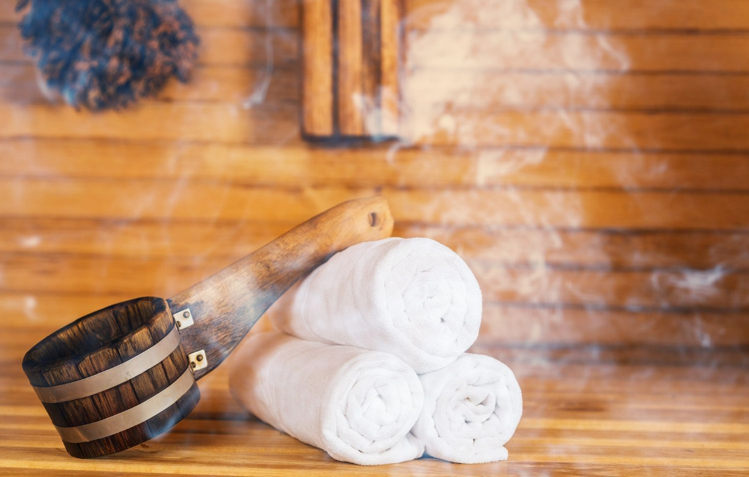White rolled towels, wooden bucket, and ladle in a sauna with steam rising.
