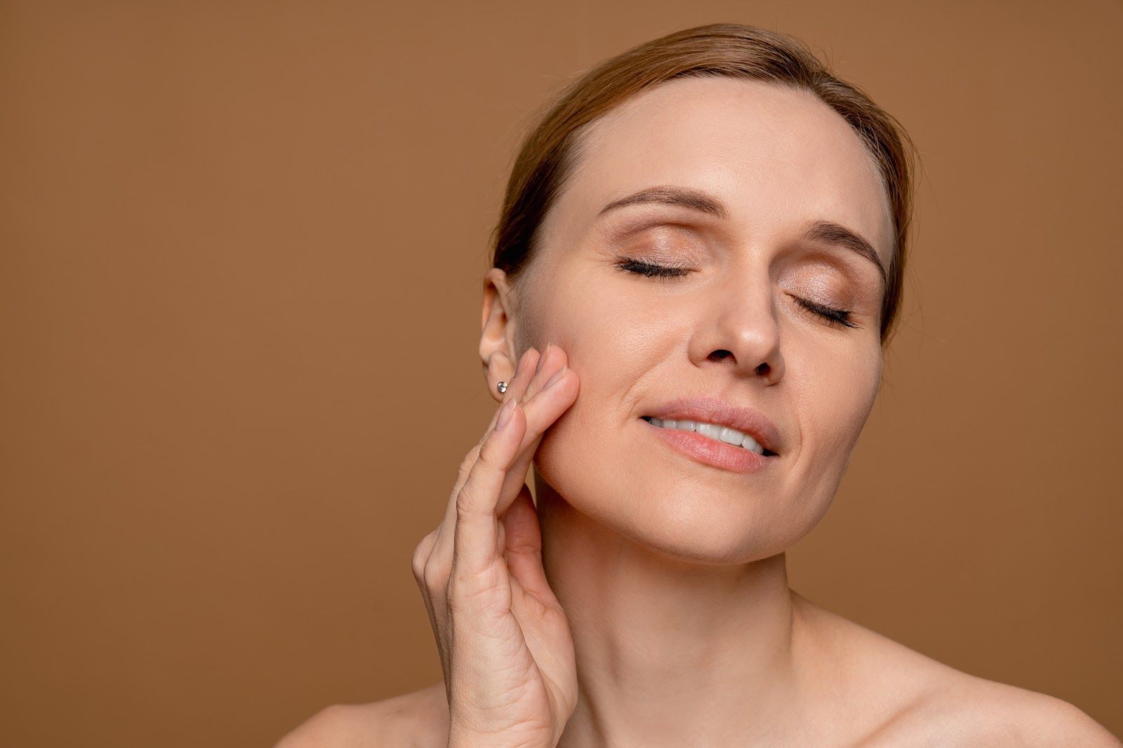 Woman with eyes closed, touching her cheek, smiling slightly against a brown background.