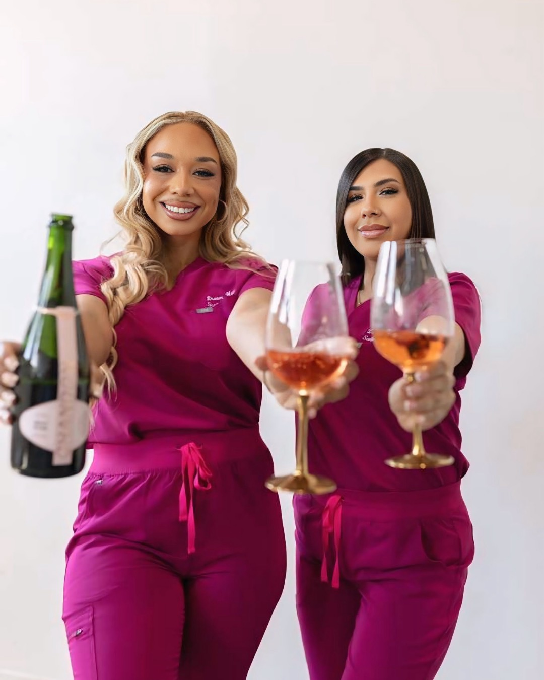 Two women in pink scrubs holding wine glasses and a bottle of sparkling wine.