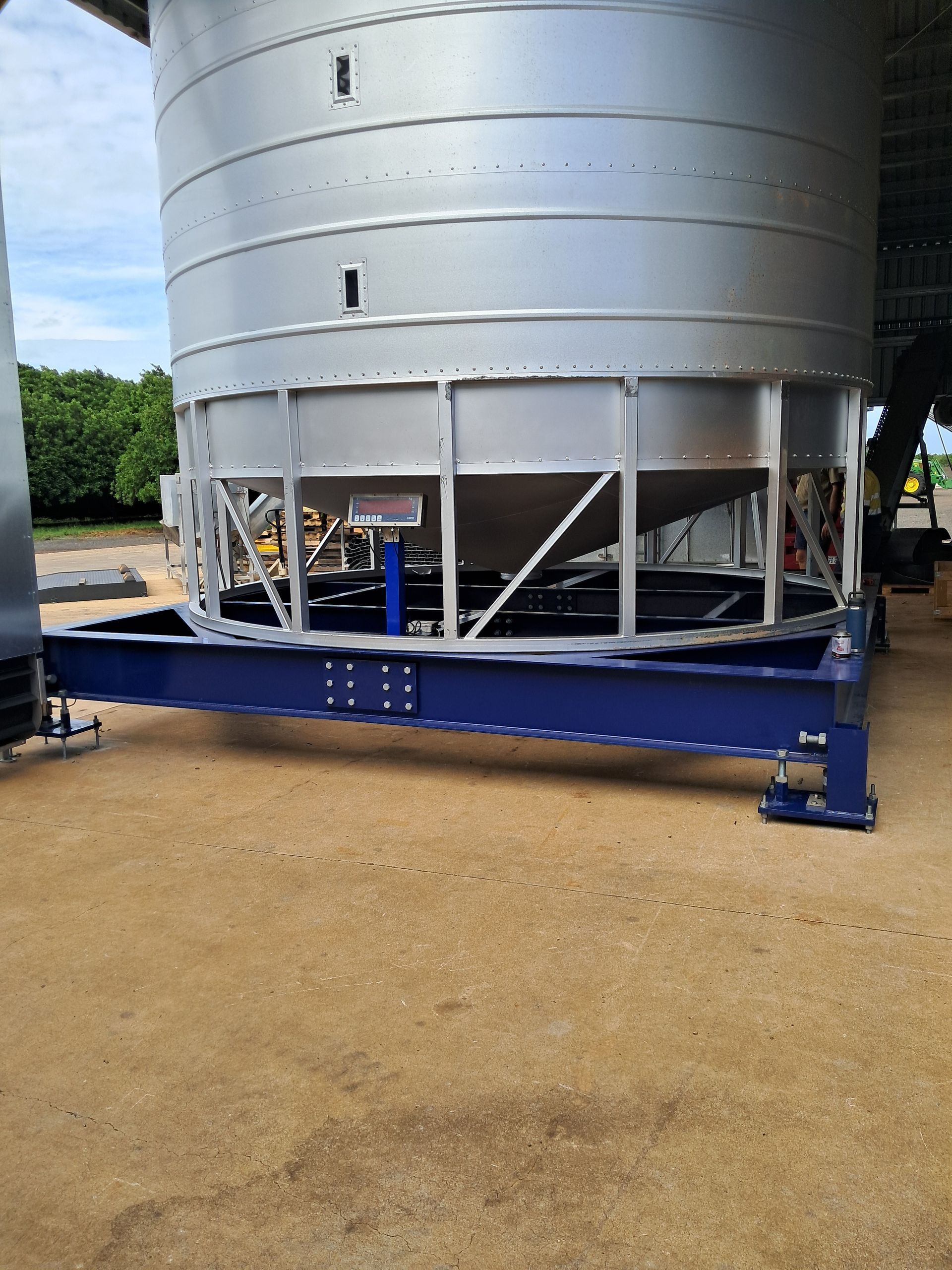 A large, silver grain silo on a blue steel frame in an outdoor industrial setting.