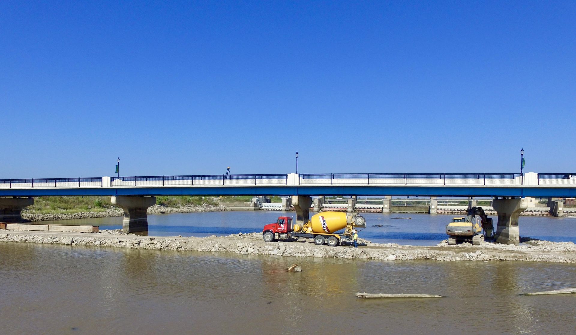 A yellow truck is driving under a bridge over a body of water.