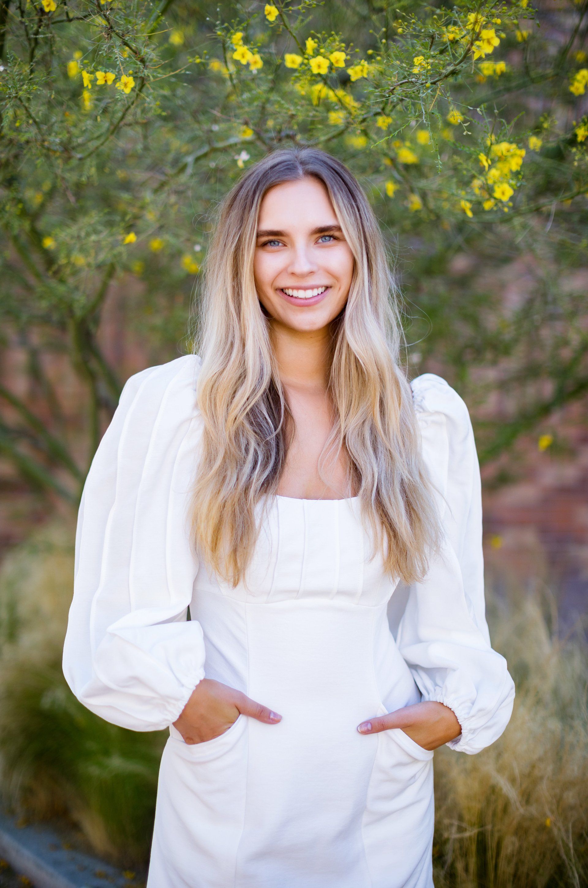 A woman in a white dress is standing in front of a tree with yellow flowers.