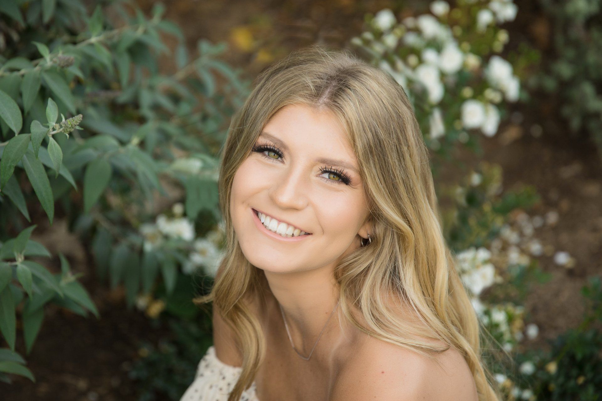 A woman is smiling in front of a bush with white flowers.