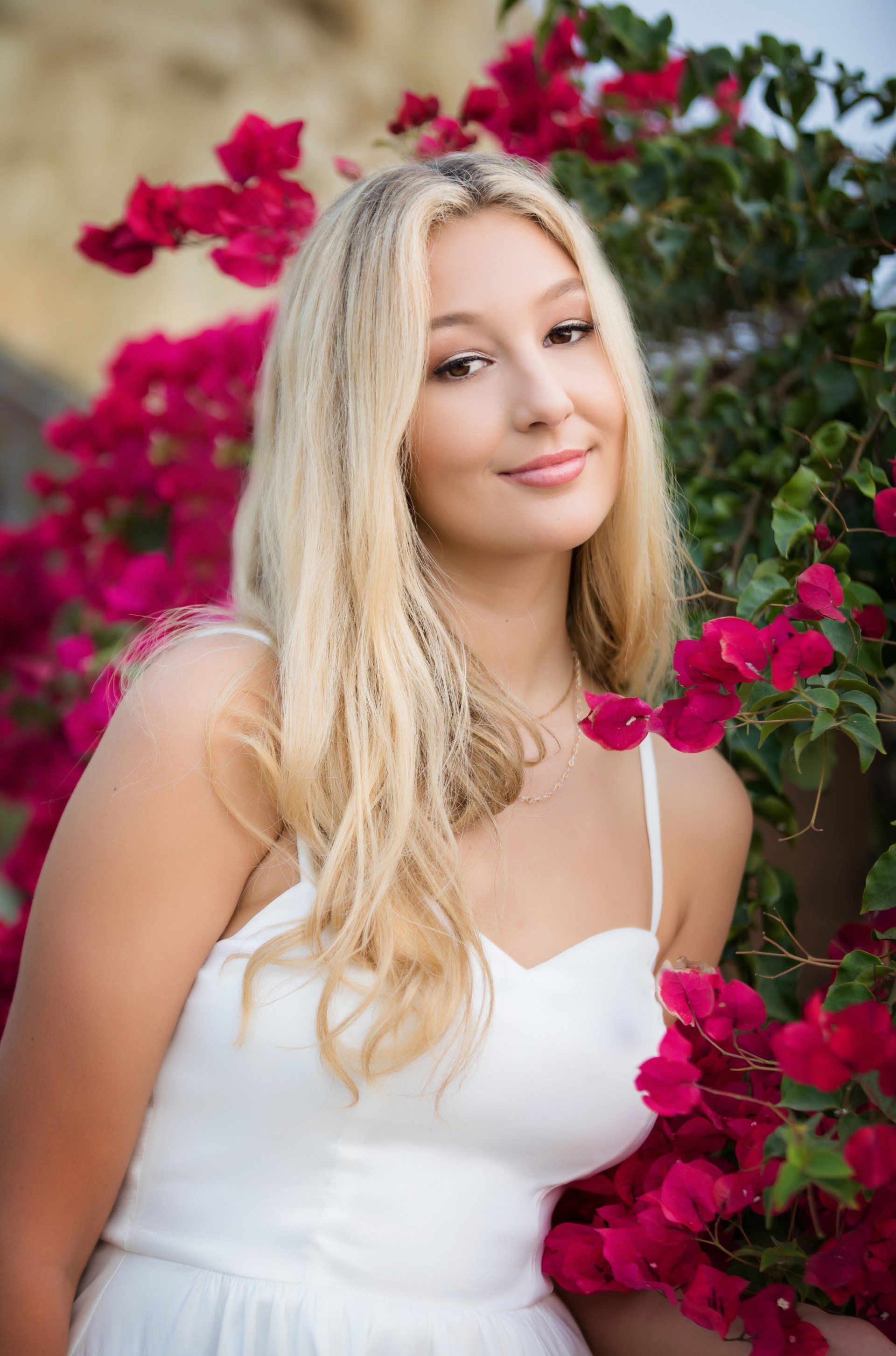 A woman in a white dress is standing in front of a bush of pink flowers.