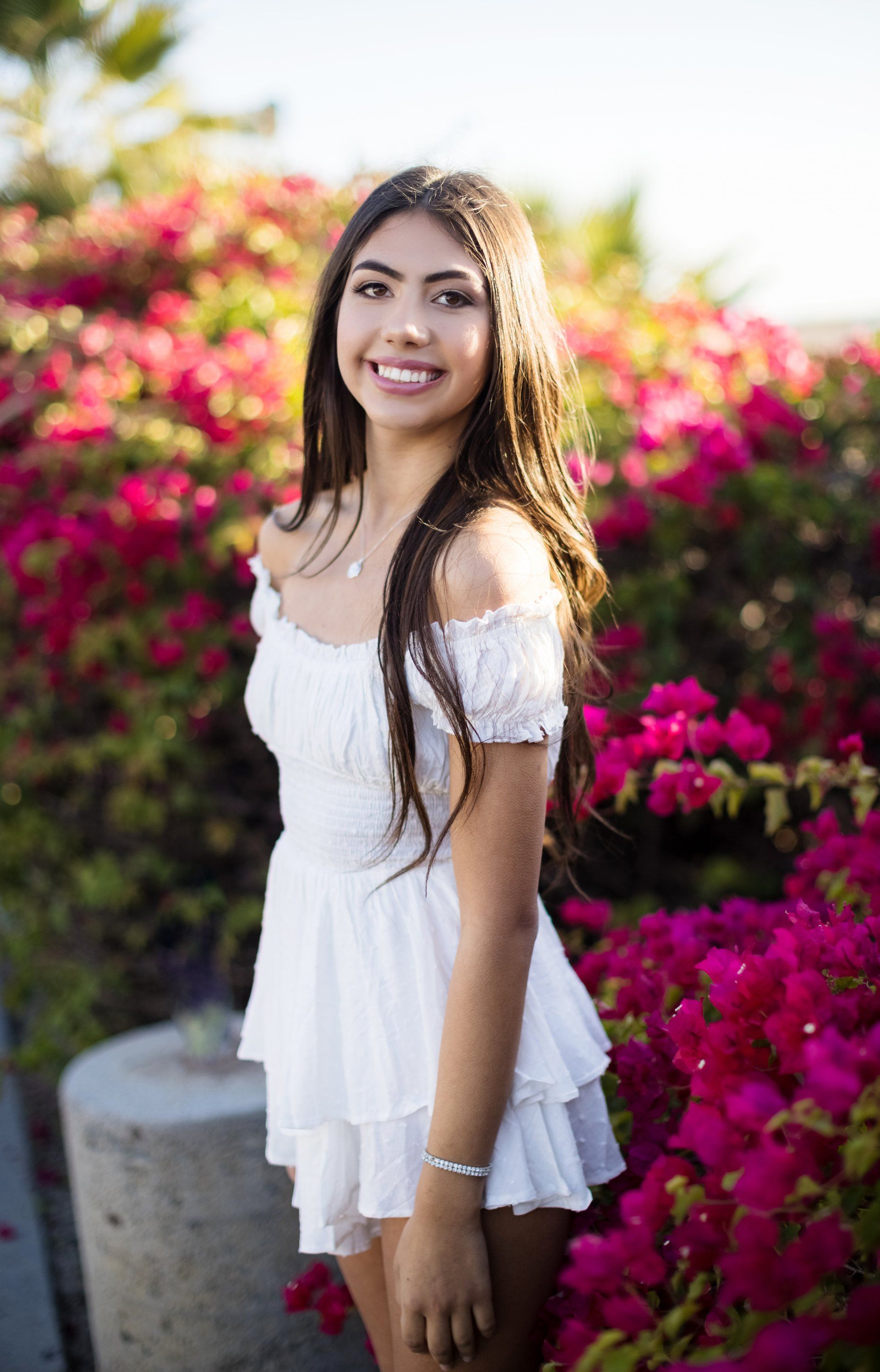 A woman in a white dress is standing in front of a bush of pink flowers.