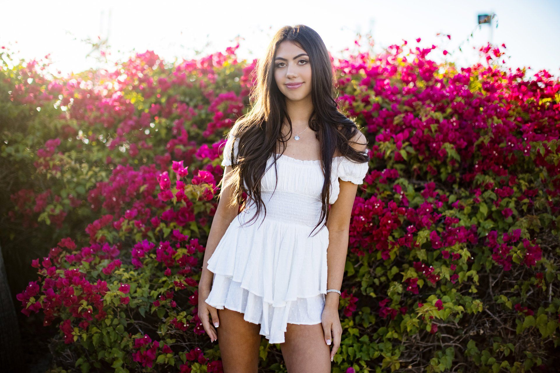 A woman in a white dress is standing in front of a bush of pink flowers.