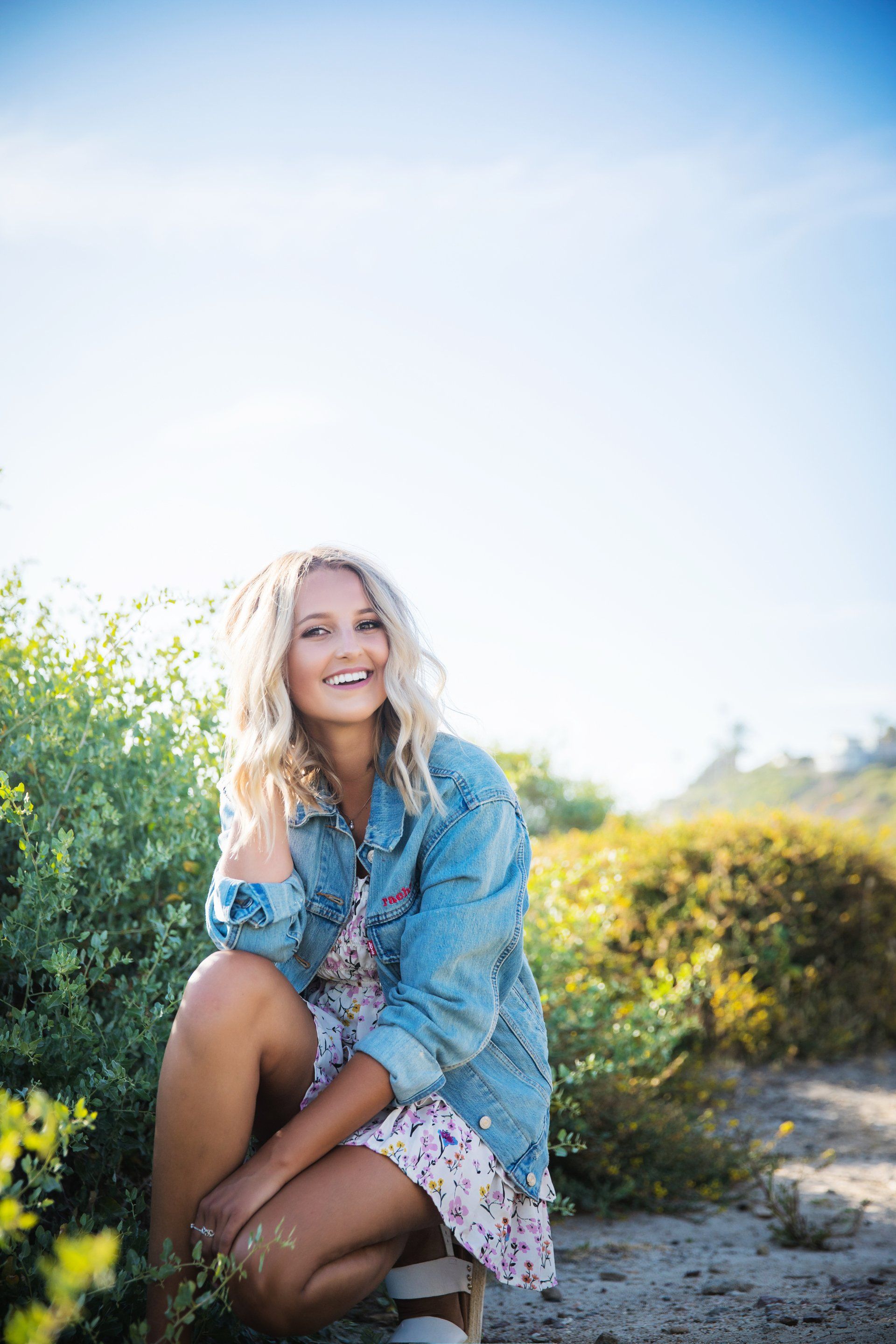 A woman in a denim jacket and floral dress is kneeling down in a field.
