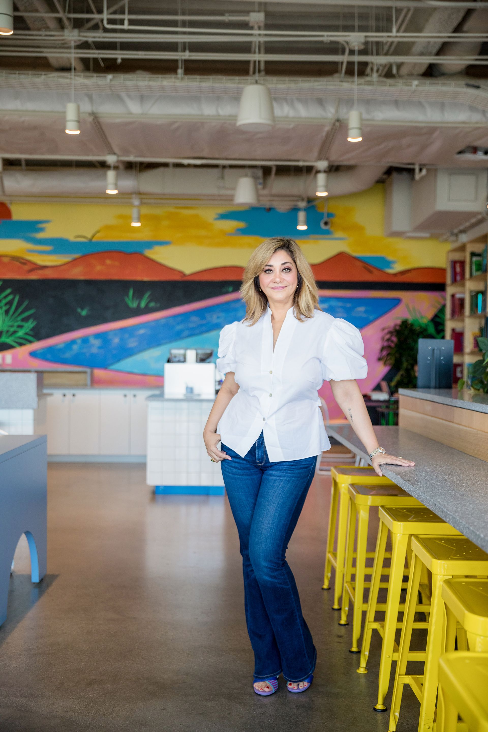 A woman in a white shirt and blue jeans is standing in a room with yellow stools.