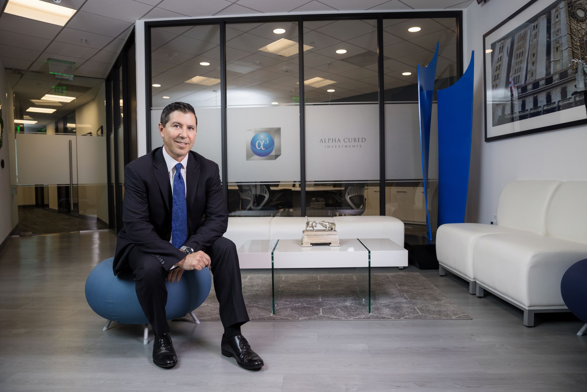 A man in a suit and tie is sitting on a blue ottoman in a waiting room.