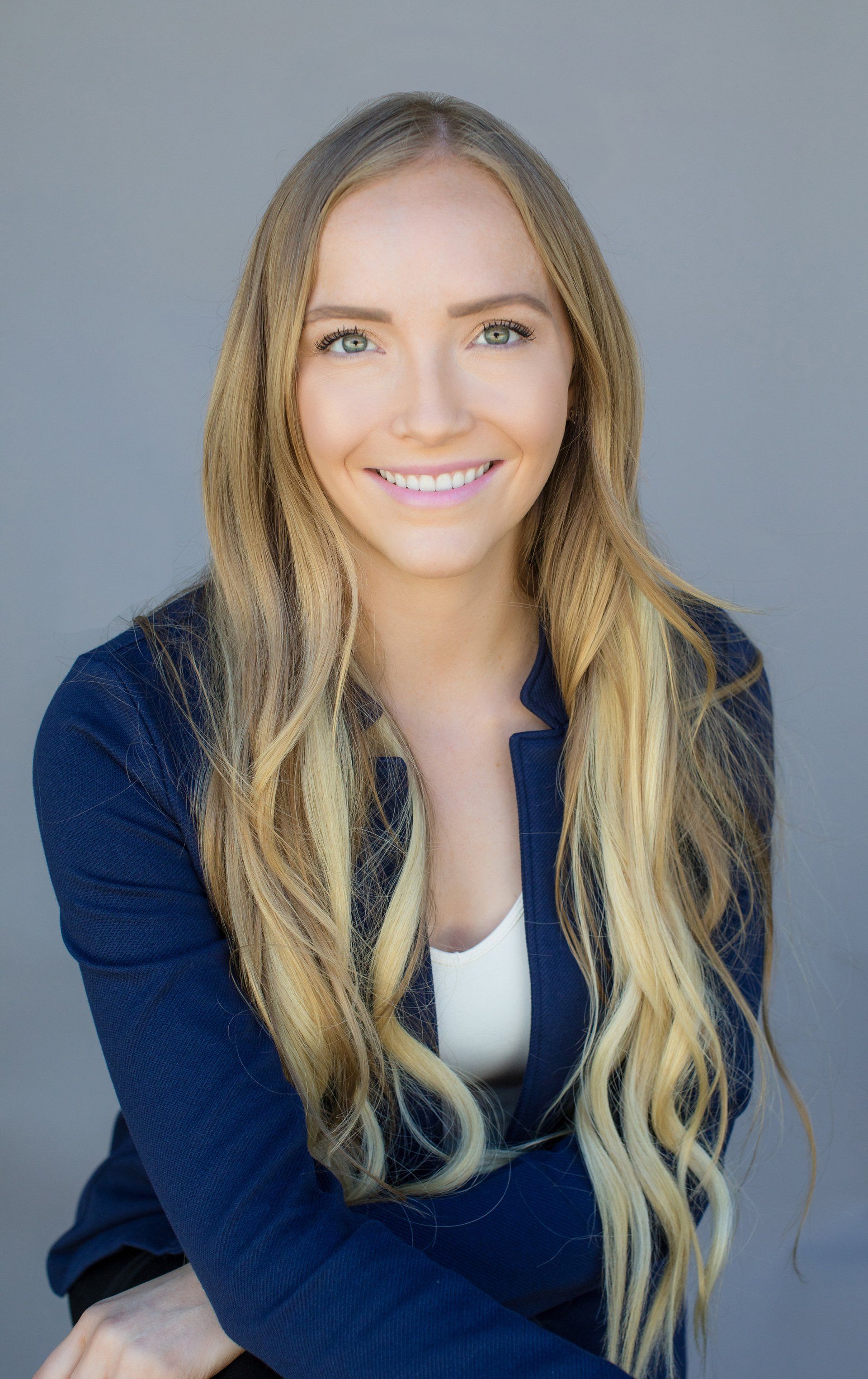 A woman with long blonde hair is wearing a blue jacket and smiling.