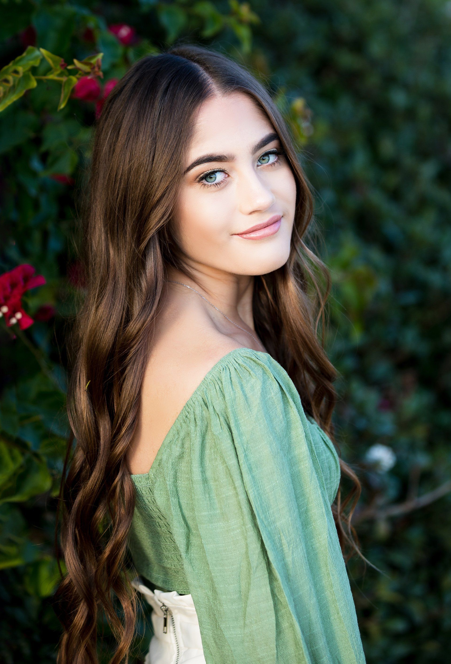 A young woman in a green dress is standing in front of a bush with red flowers.