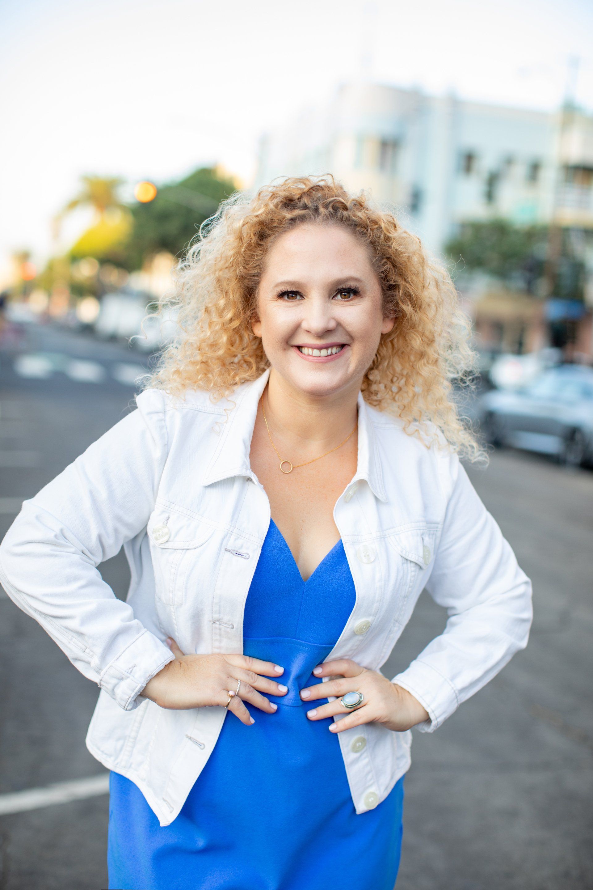 A woman in a blue dress and white jacket is standing on a city street.