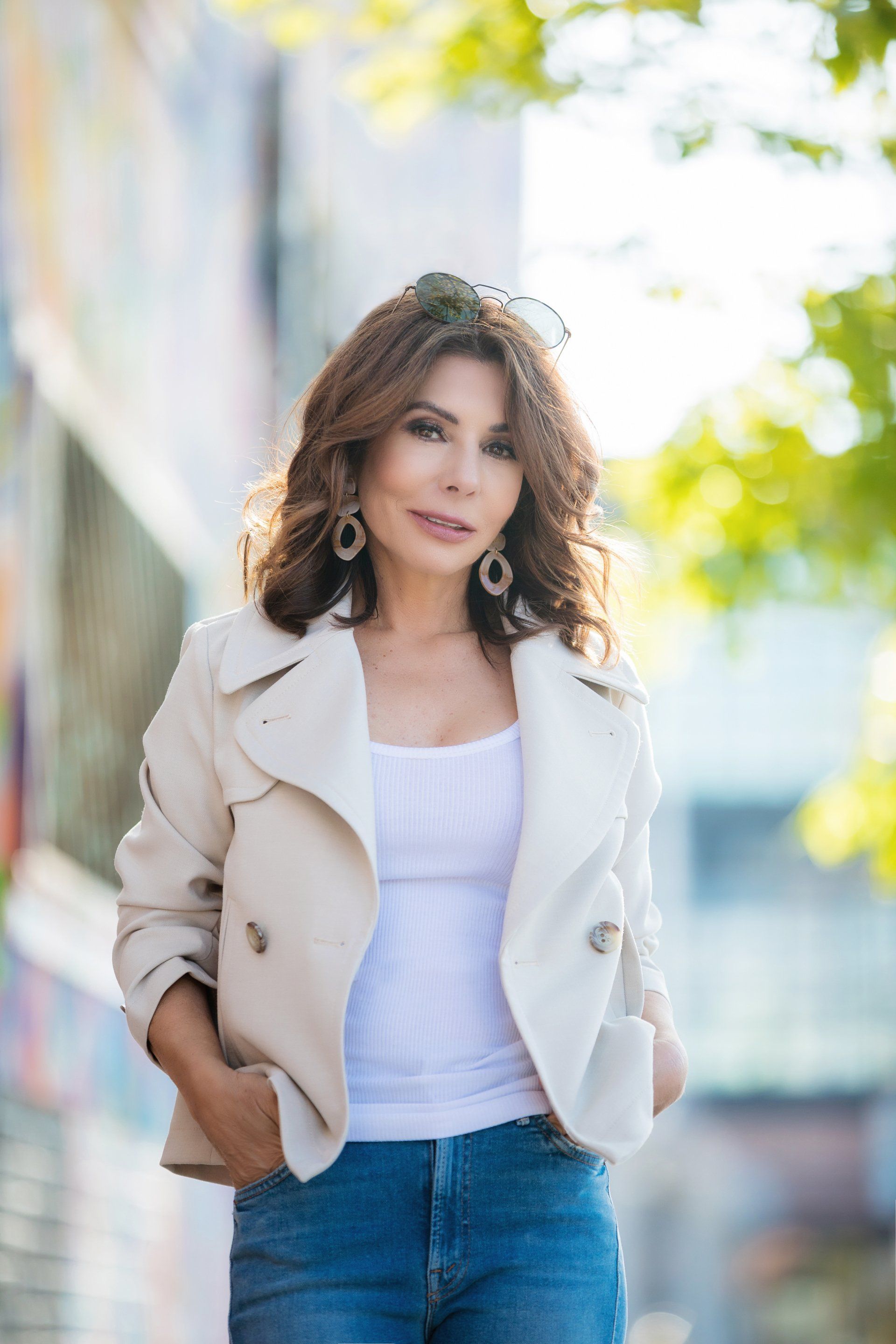 A woman wearing a white jacket and jeans is standing with her hands in her pockets.