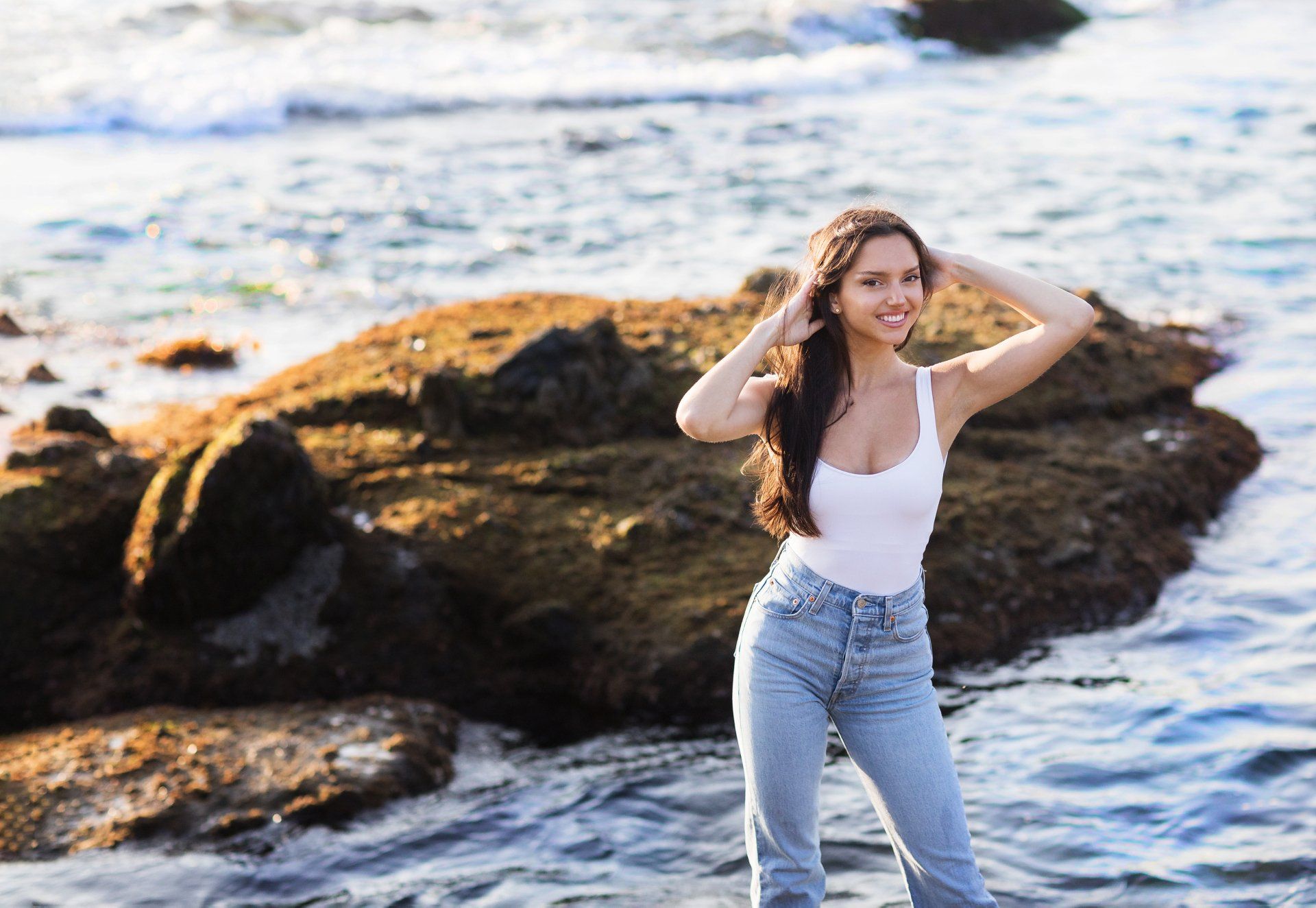 A woman in a white tank top and jeans is standing on a rocky beach near the ocean.