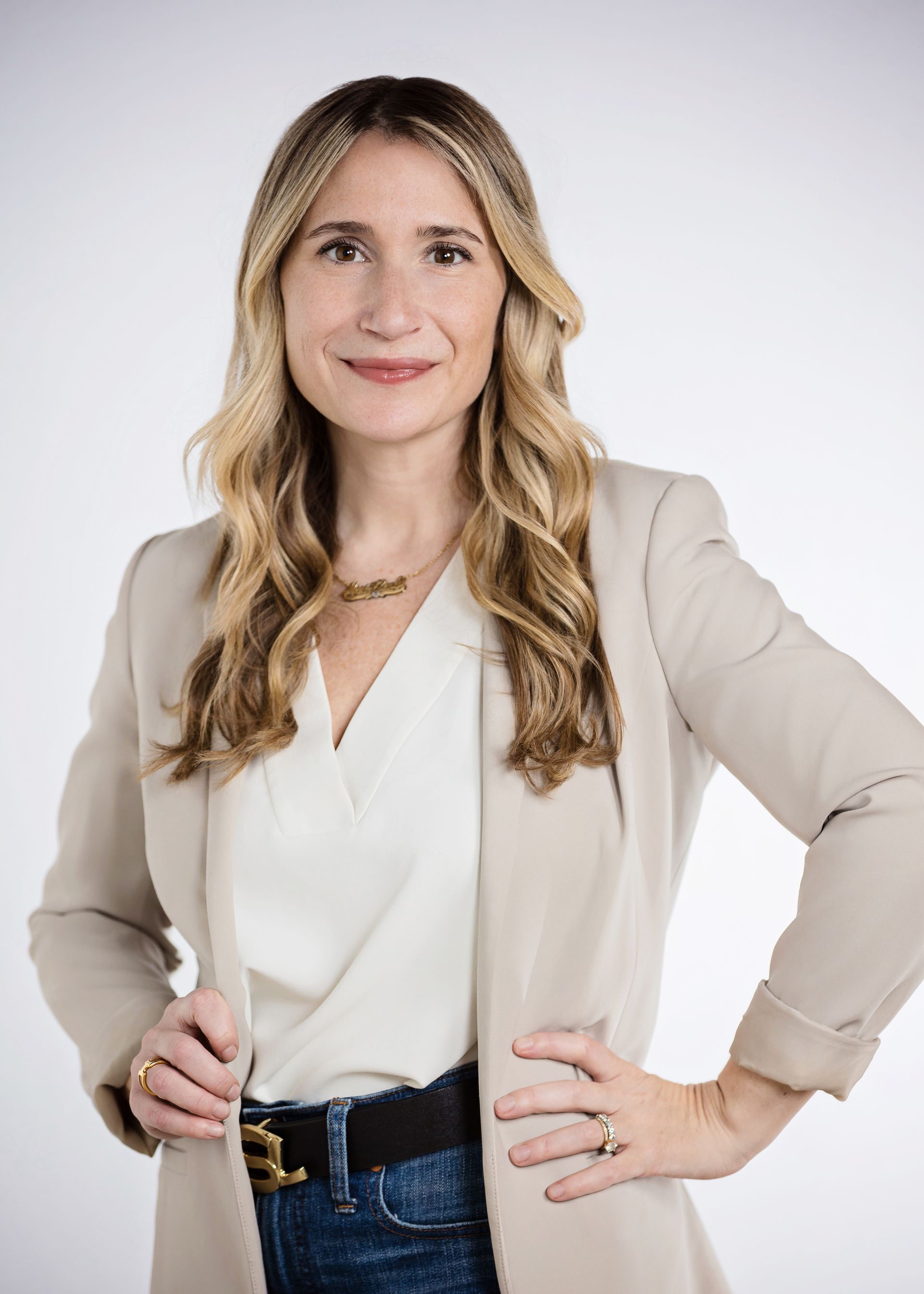 A woman in a white shirt and a tan jacket is standing with her hands on her hips.