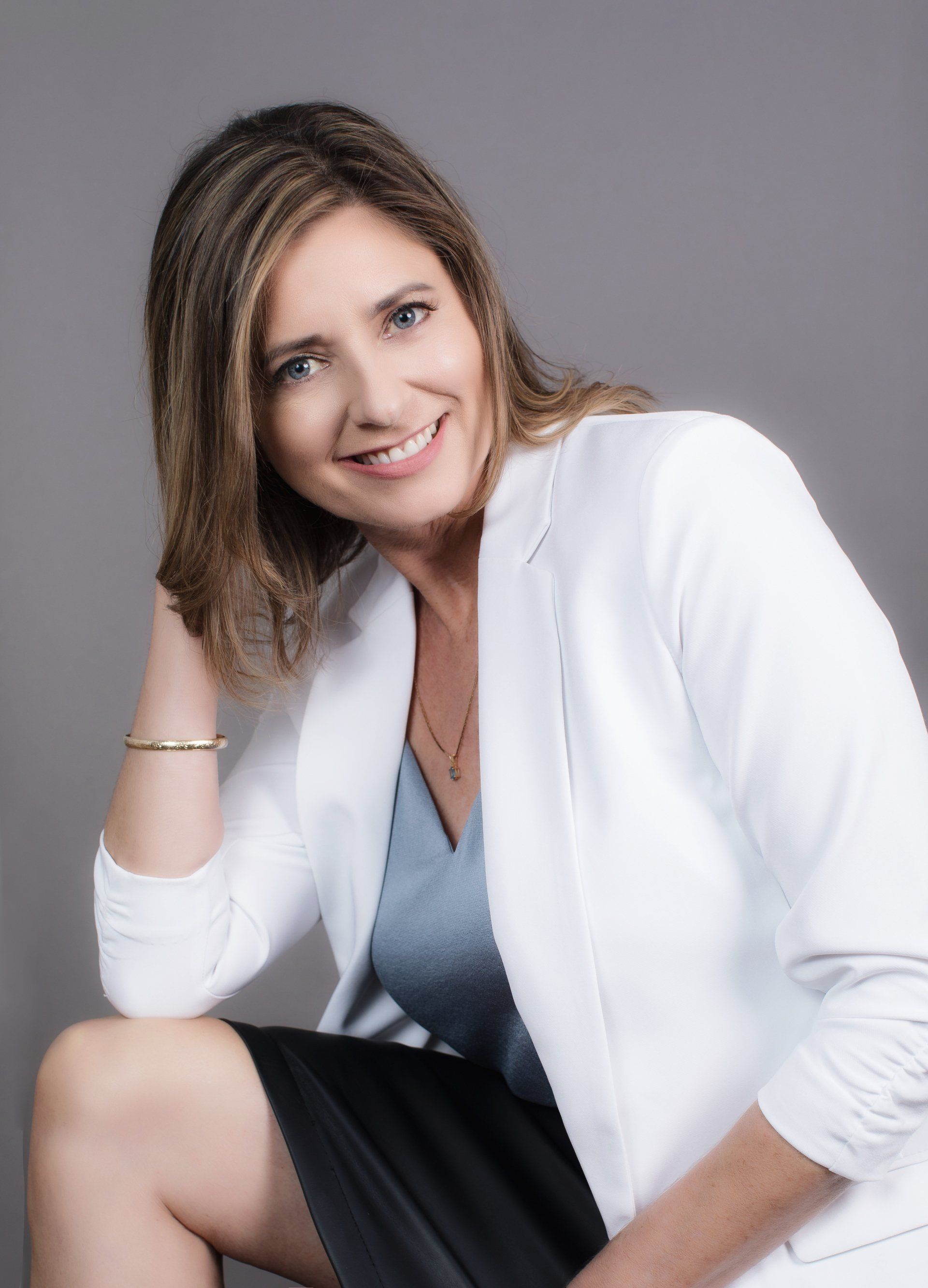 A woman in a white jacket and black skirt is sitting with her legs crossed and smiling.