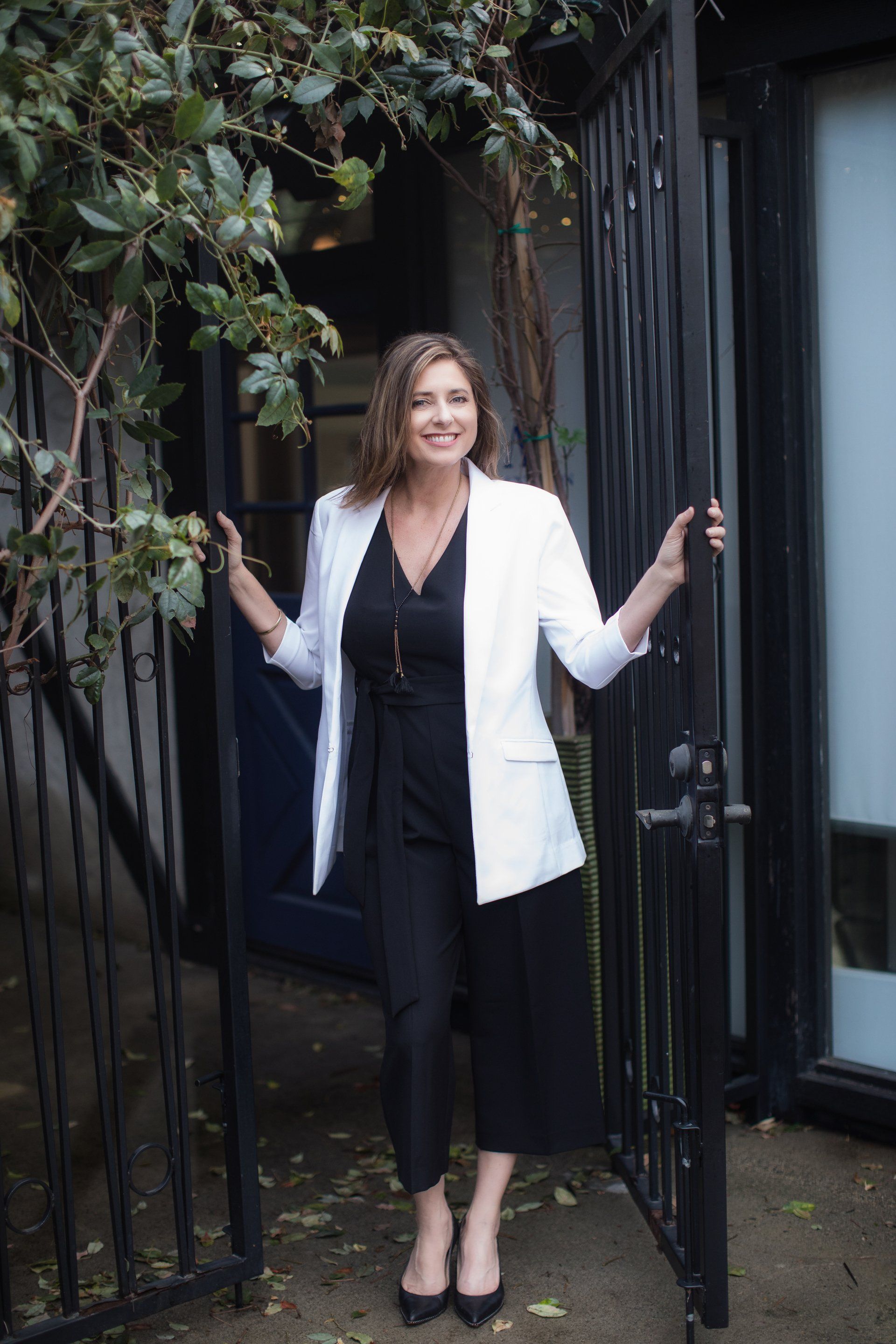 A woman in a white jacket and black jumpsuit is standing in front of a gate.